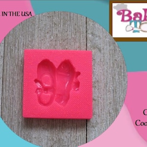 May include: A bright pink silicone mold featuring the imprint of two ballet slippers. The mold is square and sits on a wooden surface. The image also includes the text "Baker's Toybox".