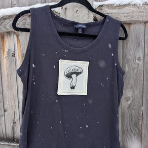 Könnte beinhalten: Dunkelmarineblaues Tanktop mit einem Pilz-Grafik-Patch. Der Patch ist ein Quadrat aus cremefarbenem Stoff mit einer Schwarz-Weiß-Illustration eines Pilzes. Das Tanktop hängt an einem schwarzen Kleiderbügel vor einem Holzzaun.