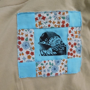 Puede incluir: Una sudadera beige con un parche cuadrado con una ilustración de un pájaro. El parche está enmarcado con tela azul claro y tela estampada floral en rojo, naranja y azul. El pájaro es negro y azul.