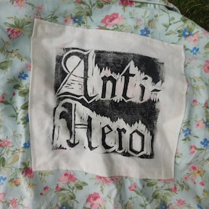 May include: A light blue shirt featuring a floral pattern of pink roses and small blue flowers. A square off-white fabric patch is sewn onto the back, displaying the black ink text Anti-Hero in a stylized, distressed gothic-inspired font.
