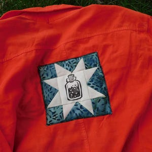 May include: Vibrant orange shirt with a square patch. The patch has a white star design and a blue patterned background. A black and white jar illustration is in the centre of the patch.