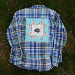 Op de afbeelding: Blauw geruit button-down shirt met een lichtblauwe vierkante patch op de rug. De patch heeft een sterontwerp met een centraal beeld van een zwarte vogel. Het shirt is gemaakt van een zacht, comfortabel materiaal.