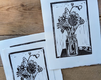 Pode incluir: Duas impressões em preto e branco de um buquê de flores em um frasco de vidro sobre uma mesa. As flores são girassóis e outras flores silvestres. As impressões são em papel branco com uma textura áspera.