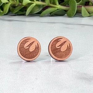 May include: A pair of wooden earrings with a Rebel Alliance symbol. The earrings are a light brown color and have a circular shape.