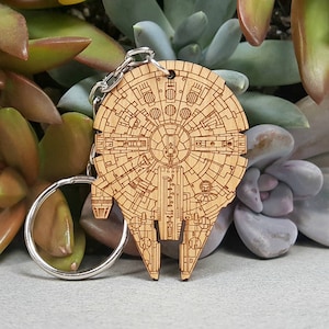 May include: A wooden keychain shaped like the Millennium Falcon from Star Wars. The keychain is detailed with the ship's design and has a silver metal ring.