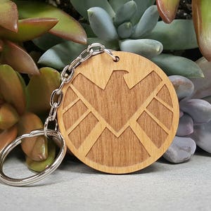 May include: A wooden keychain with a brown, circular design featuring the logo of S.H.I.E.L.D., a fictional organization from the Marvel Universe. The logo is a stylized eagle with wings spread out, surrounded by a circle with a textured wood grain.