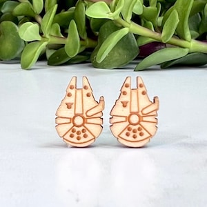 May include: A pair of wooden earrings shaped like the Millennium Falcon spaceship from Star Wars. The earrings are a light brown color and have a detailed design.