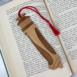 May include: A wooden bookmark with a pen-like design, featuring a light brown body and darker brown accents. A red tassel hangs from the top. The bookmark is placed on the pages of an open book.