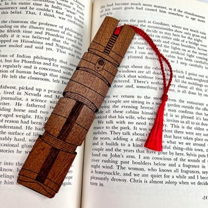 Puede incluir: Un marcap&aacute;ginas de madera con un borla roja, que presenta una ilustraci&oacute;n detallada de un sable de luz. El marcap&aacute;ginas est&aacute; sobre un libro abierto con texto sobre una pareja sentada en una caba&ntilde;a.