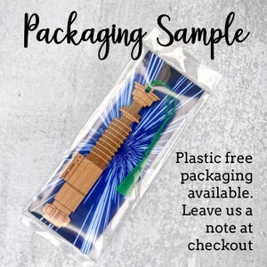 May include: A wooden lightsaber bookmark with a green tassel. The bookmark is packaged in a clear plastic sleeve with a blue and white starry sky design. The text "Plastic free packaging available. Leave us a note at checkout" is printed on the sleeve.