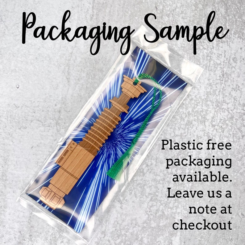 May include: A wooden lightsaber bookmark with a green tassel. The bookmark is packaged in a clear plastic sleeve with a blue and white starry sky design. The text "Plastic free packaging available. Leave us a note at checkout" is printed on the sleeve.