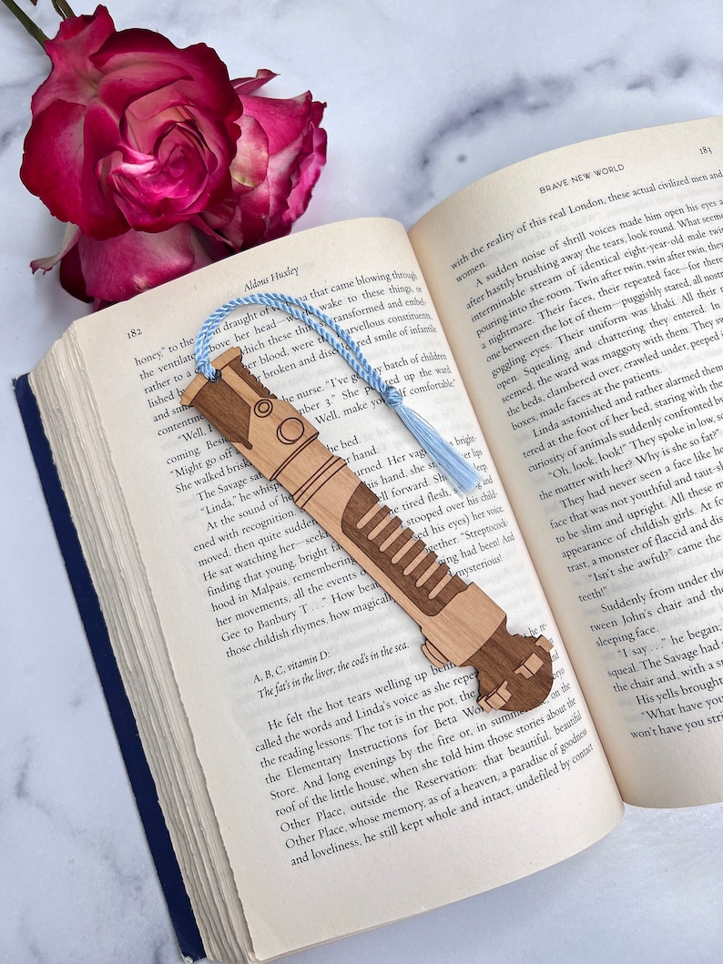 May include: A wooden bookmark shaped like a lightsaber from the Star Wars franchise. The bookmark is brown and has a light brown handle. The bookmark is resting on an open book with the text "Brave New World" visible.