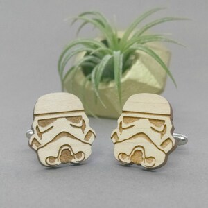 May include: A pair of wooden cufflinks featuring a laser-engraved design of a Star Wars Stormtrooper helmet. The cufflinks are a light brown color and have a silver metal backing.
