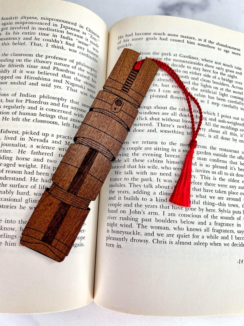 Puede incluir: Un marcap&aacute;ginas de madera con un borla roja, que presenta una ilustraci&oacute;n detallada de un sable de luz. El marcap&aacute;ginas est&aacute; sobre un libro abierto con texto sobre una pareja sentada en una caba&ntilde;a.