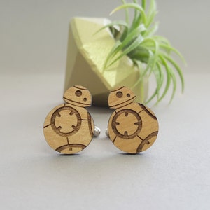 May include: A pair of wooden cufflinks featuring a laser-engraved design of a spherical robot with a circular pattern. The cufflinks are a light brown color.