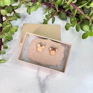 Peut inclure: Une paire de boucles d'oreilles en bois dans une petite bo&icirc;te brune. Les boucles d'oreilles sont en forme de t&ecirc;tes de Mickey et Minnie Mouse. La boucle d'oreille Mickey Mouse est en bois uni, tandis que la boucle d'oreille Minnie Mouse a un petit n&oelig;ud rouge &agrave; pois blancs.