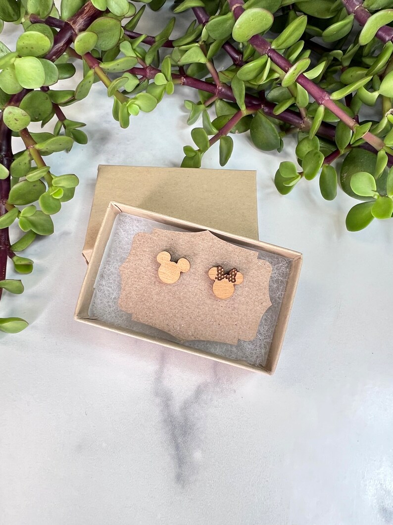Peut inclure: Une paire de boucles d'oreilles en bois dans une petite bo&icirc;te brune. Les boucles d'oreilles sont en forme de t&ecirc;tes de Mickey et Minnie Mouse. La boucle d'oreille Mickey Mouse est en bois uni, tandis que la boucle d'oreille Minnie Mouse a un petit n&oelig;ud rouge &agrave; pois blancs.