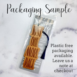 Puede incluir: Un marcap&aacute;ginas de madera con el texto "Just One More Page" y un borla azul. El marcap&aacute;ginas est&aacute; empaquetado en una bolsa de pl&aacute;stico transparente con una tarjeta blanca que dice "Embalaje sin pl&aacute;stico disponible. D&eacute;jenos una nota en la caja."