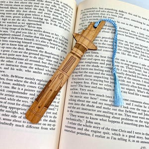 Op de afbeelding: Een houten bladwijzer in de vorm van een lightsaber, met een blauwe kwast. De bladwijzer ligt op een open boek met tekst over een reis naar Montana.