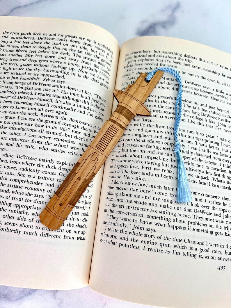 Op de afbeelding: Een houten bladwijzer in de vorm van een lightsaber, met een blauwe kwast. De bladwijzer ligt op een open boek met tekst over een reis naar Montana.