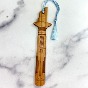 Op de afbeelding: Een houten bladwijzer in de vorm van een lightsaber met een blauwe kwast. De bladwijzer is gemaakt van lichtbruin hout en heeft een gedetailleerd ontwerp.