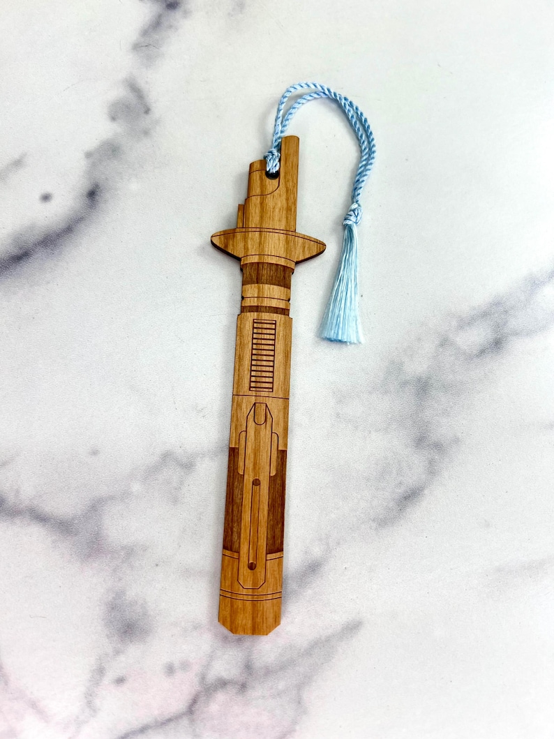 Op de afbeelding: Een houten bladwijzer in de vorm van een lightsaber met een blauwe kwast. De bladwijzer is gemaakt van lichtbruin hout en heeft een gedetailleerd ontwerp.