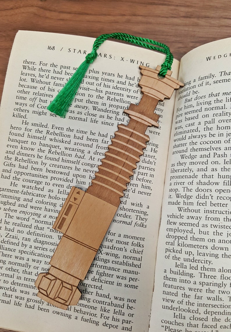 May include: A wooden bookmark shaped like an X-Wing fighter from Star Wars. The bookmark is painted in shades of brown and has a green tassel.