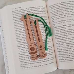 May include: Two wooden bookmarks shaped like lightsabers with green tassels. The bookmarks are engraved with details and have a wood grain finish.