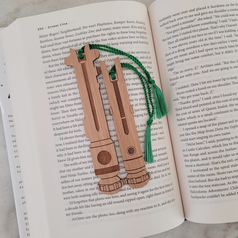 May include: Two wooden bookmarks shaped like lightsabers with green tassels. The bookmarks are engraved with details and have a wood grain finish.