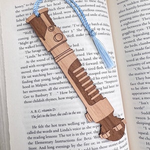 May include: A wooden bookmark shaped like a futuristic weapon, with a blue tassel. The bookmark is resting on an open book with text about Aldous Huxley's novel "Brave New World".