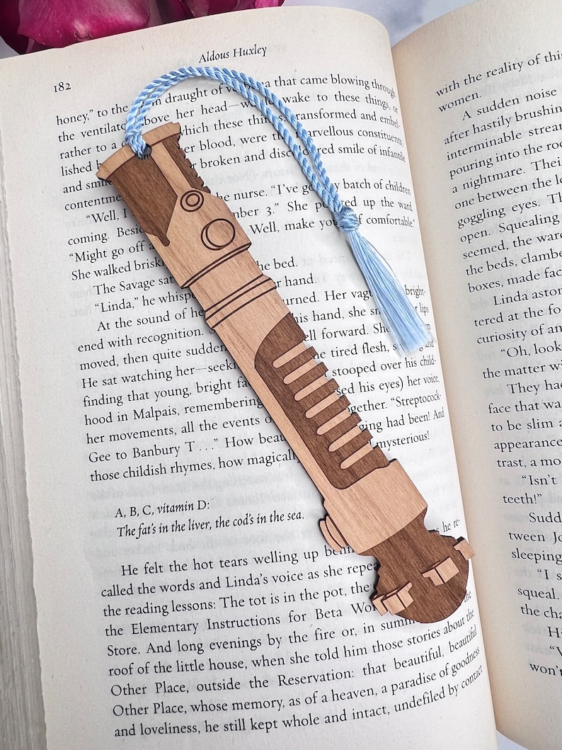 May include: A wooden bookmark shaped like a futuristic weapon, with a blue tassel. The bookmark is resting on an open book with text about Aldous Huxley's novel "Brave New World".