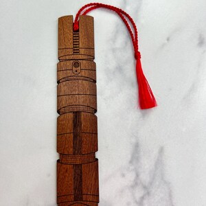 Puede incluir: Un marcap&aacute;ginas de madera con forma de sable de luz con un borla roja. El marcap&aacute;ginas est&aacute; hecho de madera oscura con un patr&oacute;n de veta.