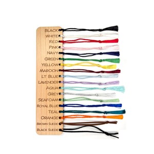 May include: A wooden swatch card with 18 different colored cords attached. The cords are labeled with the following colors: black, white, red, pink, navy, green, yellow, maroon, light blue, lavender, aqua, grey, seafoam, royal blue, teal, orange, brown suede, and black suede.