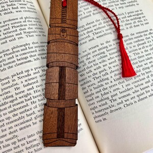 Puede incluir: Un marcap&aacute;ginas de madera con un dise&ntilde;o detallado de un faro. El marcap&aacute;ginas es marr&oacute;n con un borde marr&oacute;n oscuro y est&aacute; apoyado en un libro.