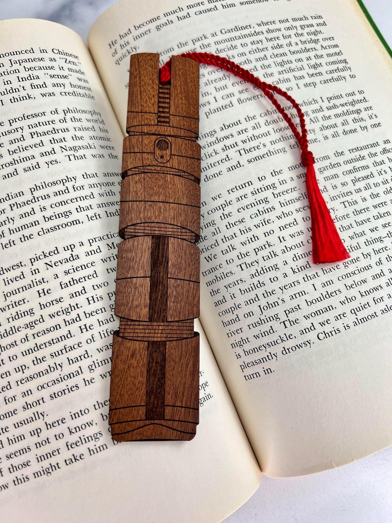 Puede incluir: Un marcap&aacute;ginas de madera con un dise&ntilde;o detallado de un faro. El marcap&aacute;ginas es marr&oacute;n con un borde marr&oacute;n oscuro y est&aacute; apoyado en un libro.