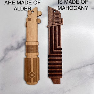 Puede incluir: Dos r&eacute;plicas de sable de luz de madera. El sable de luz de la izquierda est&aacute; hecho de madera de aliso y tiene el texto "OTHER LIGHTSABERS ARE MADE OF ALDER". El sable de luz de la derecha est&aacute; hecho de madera de caoba y tiene el texto "DARKSABER IS MADE OF MAHOGANY".