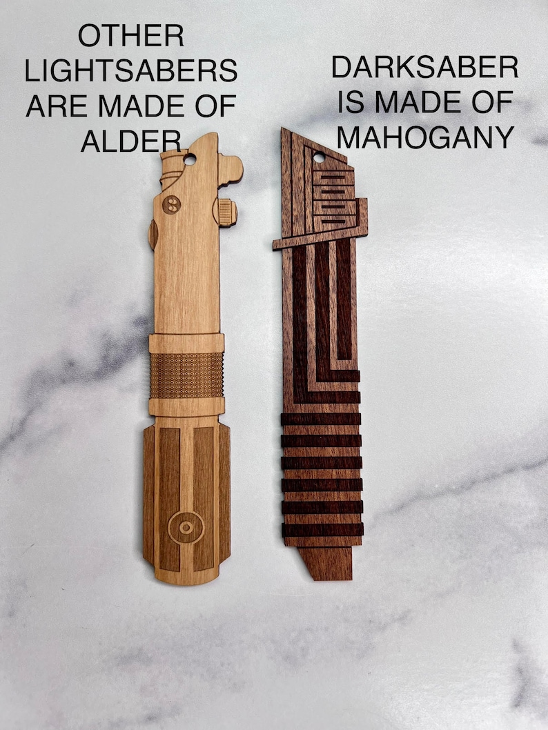 Puede incluir: Dos r&eacute;plicas de sable de luz de madera. El sable de luz de la izquierda est&aacute; hecho de madera de aliso y tiene el texto "OTHER LIGHTSABERS ARE MADE OF ALDER". El sable de luz de la derecha est&aacute; hecho de madera de caoba y tiene el texto "DARKSABER IS MADE OF MAHOGANY".