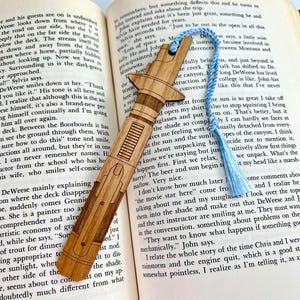 Op de afbeelding: Een houten bladwijzer in de vorm van een lightsaber, met een blauwe kwast. De bladwijzer ligt op een open boek met tekst over een reis naar Minnesota. De tekst op de bladwijzer luidt "Just to be out in the open in all this space."