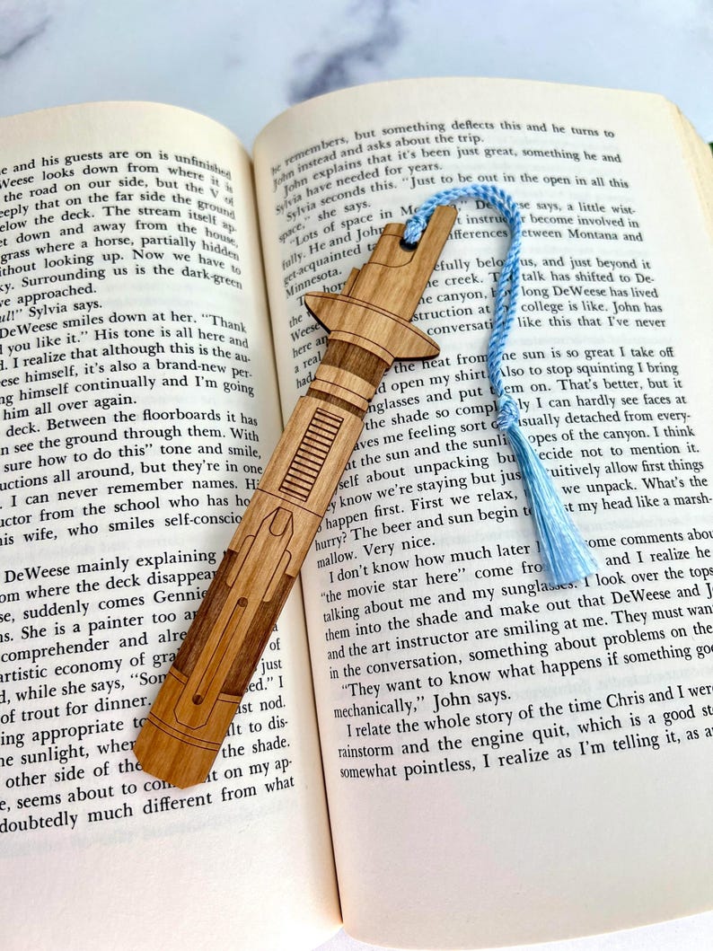 Op de afbeelding: Een houten bladwijzer in de vorm van een lightsaber, met een blauwe kwast. De bladwijzer ligt op een open boek met tekst over een reis naar Minnesota. De tekst op de bladwijzer luidt "Just to be out in the open in all this space."