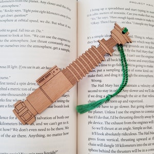 May include: A wooden bookmark shaped like a lightsaber with a green tassel. The bookmark is resting on an open book with text about space travel and a spaceship.
