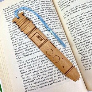 May include: Wooden bookmark with a light brown finish, featuring engraved geometric designs and a blue tassel. The bookmark is placed in an open book with text visible on the pages. A unique gift for book lovers.