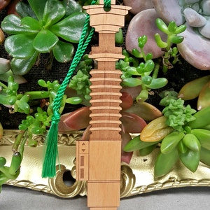 May include: A wooden lightsaber bookmark with a green tassel. The bookmark is light brown and has a detailed design.