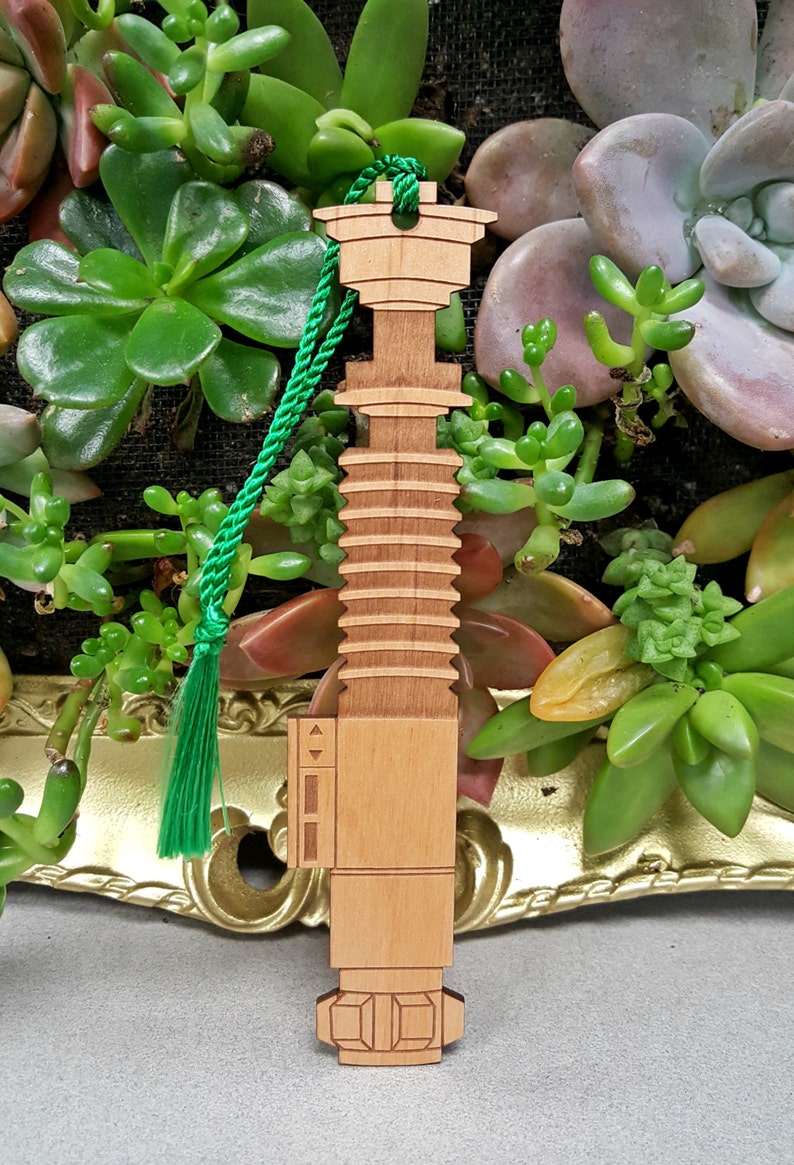 May include: A wooden lightsaber bookmark with a green tassel. The bookmark is light brown and has a detailed design.