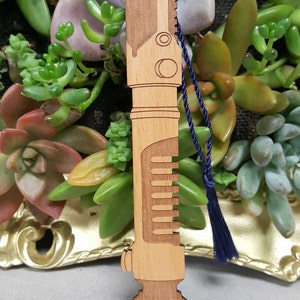 May include: A wooden lightsaber bookmark with a blue tassel. The bookmark is light brown and has a detailed design.