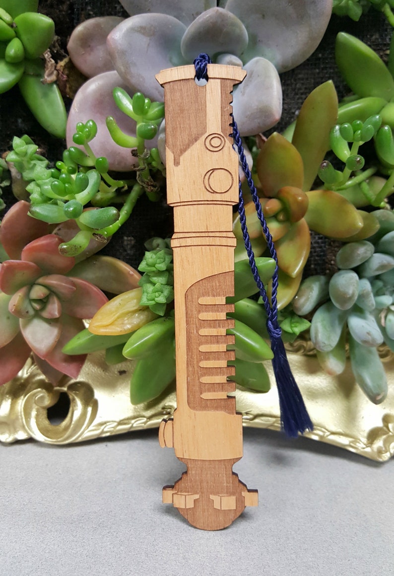 May include: A wooden lightsaber bookmark with a blue tassel. The bookmark is light brown and has a detailed design.