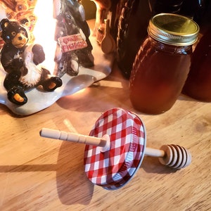 May include: A red and white gingham lid with a wooden honey dipper attached to it. The dipper has a wooden handle and a spiral-shaped honey dipper. The lid is sitting on a wooden surface.