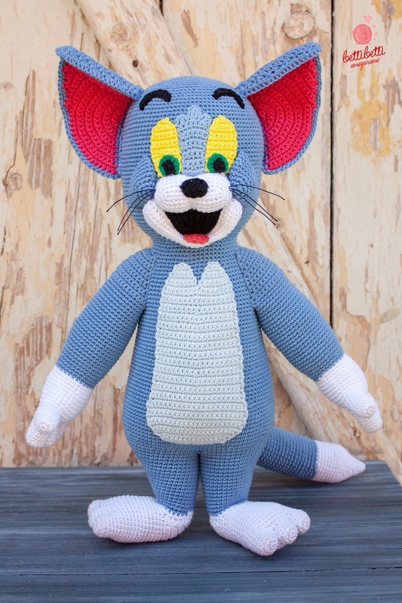 May include: A crocheted Tom the Cat plush toy with blue fur, white paws, and red ears. The toy has yellow eyes, a green nose, and a black mouth.