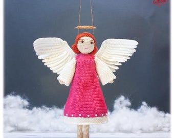 May include: A crocheted angel ornament featuring a bright pink dress with a white hem and small white beads, large white wings, orange hair, and a gold halo suspended by a thin thread. The figure has a light beige face and legs.