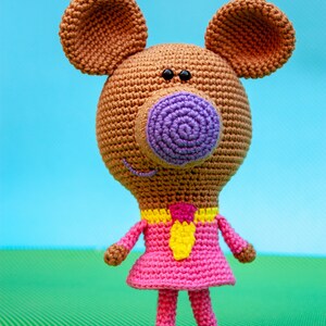 May include: A crocheted brown bear wearing a pink dress with a yellow tie. The bear has a purple nose and black eyes.