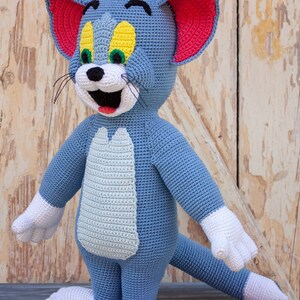 May include: A crocheted Tom cat plush toy with blue fur, white paws, and a red and white face. The toy is standing on a wooden surface.
